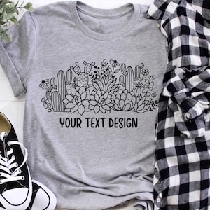 May include: A gray t-shirt with a black and white graphic of a cactus and succulent garden. The text "YOUR TEXT DESIGN" is printed below the graphic.