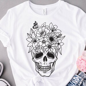 May include: A white t-shirt with a black and white illustration of a skull with flowers on top. The skull has a floral crown with a butterfly.