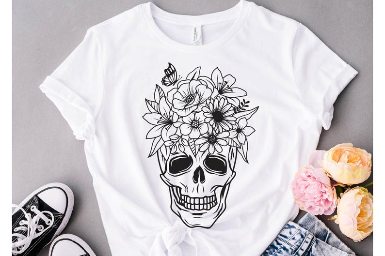 May include: A white t-shirt with a black and white illustration of a skull with flowers on top. The skull has a floral crown with a butterfly.