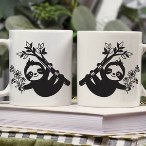 May include: Two white ceramic mugs with black sloth designs. Each sloth is hanging from a branch with flowers and butterflies. The mugs are on a stack of books.