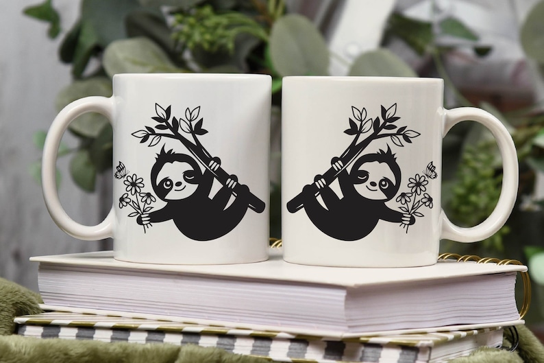 May include: Two white ceramic mugs with black sloth designs. Each sloth is hanging from a branch with flowers and butterflies. The mugs are on a stack of books.