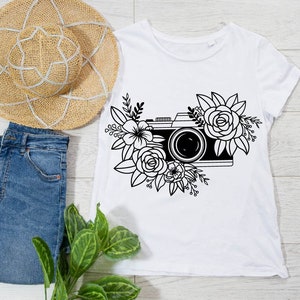 K&ouml;nnte beinhalten: Ein wei&szlig;es T-Shirt mit einer schwarz-wei&szlig;en Illustration einer Kamera, die von Blumen umgeben ist.
