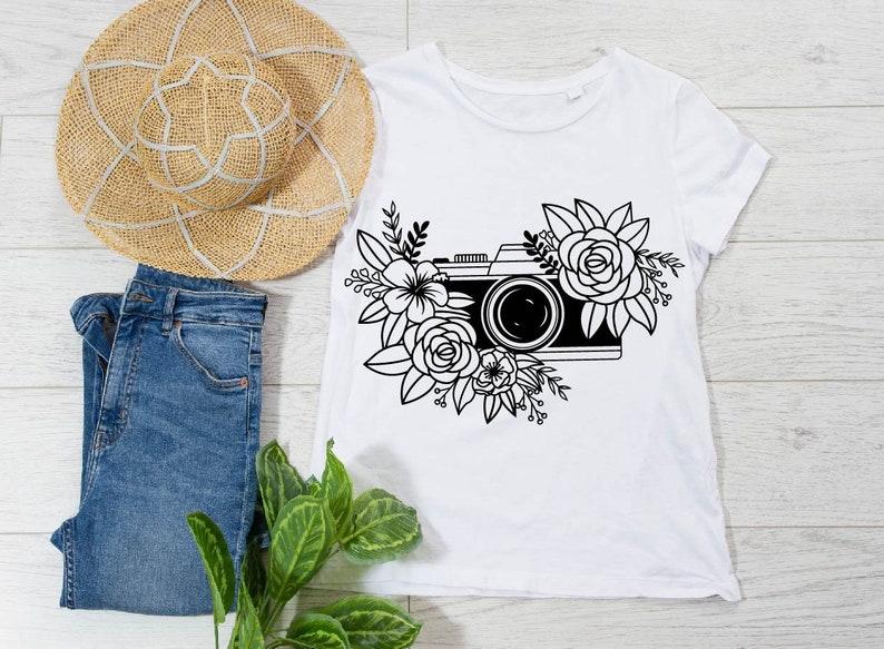 K&ouml;nnte beinhalten: Ein wei&szlig;es T-Shirt mit einer schwarz-wei&szlig;en Illustration einer Kamera, die von Blumen umgeben ist.