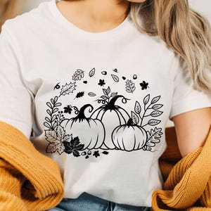 May include: White t-shirt with a black and white graphic design of three pumpkins with leaves and vines. The design is centered on the shirt.