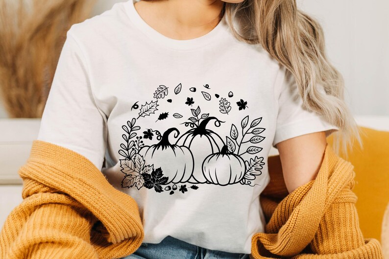 May include: White t-shirt with a black and white graphic design of three pumpkins with leaves and vines. The design is centered on the shirt.