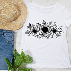 May include: White t-shirt with a black floral sunflower design. The shirt is styled with blue denim jeans and a straw sun hat. The floral design is a prominent feature on the front of the tee.