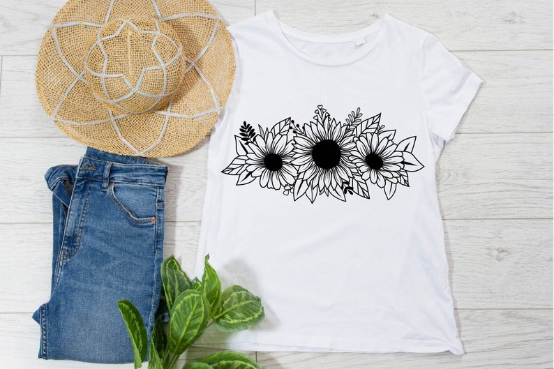 May include: White t-shirt with a black floral sunflower design. The shirt is styled with blue denim jeans and a straw sun hat. The floral design is a prominent feature on the front of the tee.