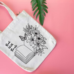 May include: White canvas tote bag with black text that reads "Book With Florals" and a black line drawing of an open book with flowers and a butterfly.