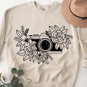 K&ouml;nnte beinhalten: Ein beiger Pullover mit einer schwarz-wei&szlig;en Illustration einer Kamera, die von Blumen umgeben ist.