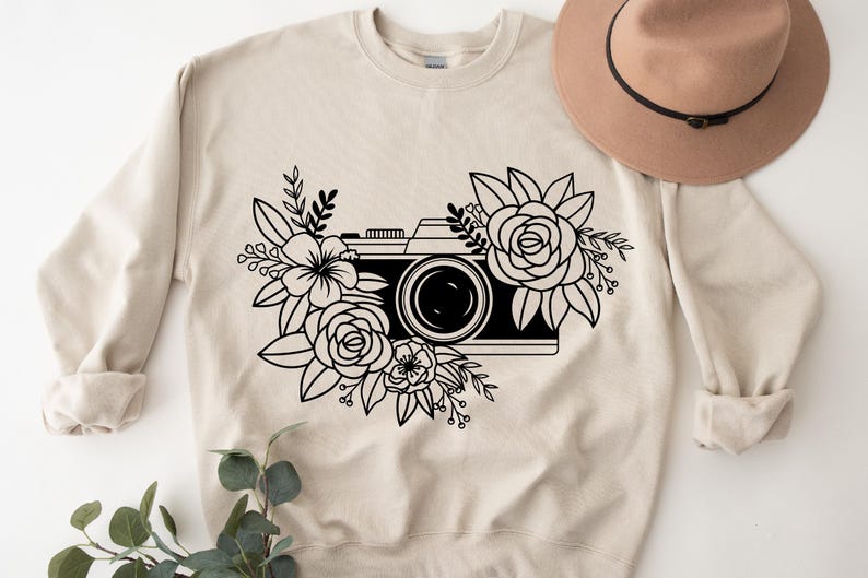 K&ouml;nnte beinhalten: Ein beiger Pullover mit einer schwarz-wei&szlig;en Illustration einer Kamera, die von Blumen umgeben ist.