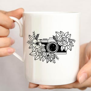 K&ouml;nnte beinhalten: Wei&szlig;e Keramiktasse mit einer Schwarzwei&szlig;illustration einer Kamera, die von Blumen umgeben ist.