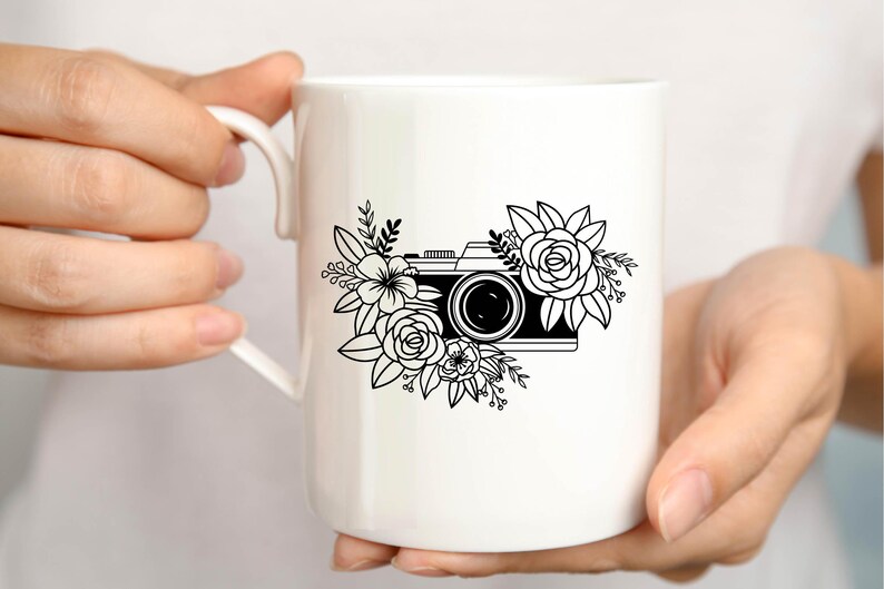 K&ouml;nnte beinhalten: Wei&szlig;e Keramiktasse mit einer Schwarzwei&szlig;illustration einer Kamera, die von Blumen umgeben ist.