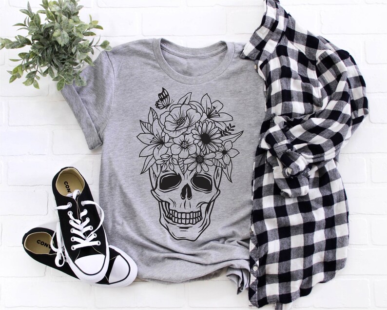 May include: A gray t-shirt with a black and white graphic of a skull with flowers on top. The skull has a floral crown with a butterfly.