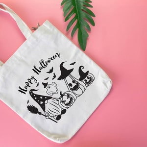 May include: White canvas tote bag with black text that says "Happy Halloween". The bag features a black and white illustration of a gnome wearing a witch hat and holding a broom, with a ghost and pumpkins behind it.