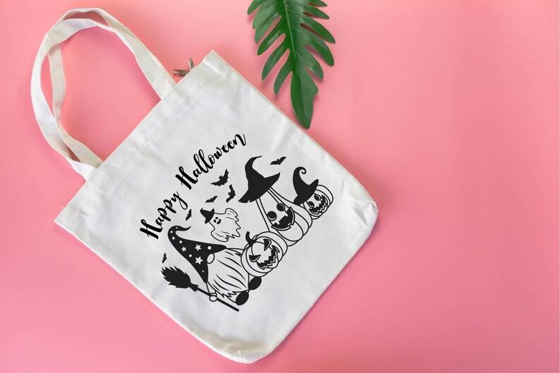 May include: White canvas tote bag with black text that says "Happy Halloween". The bag features a black and white illustration of a gnome wearing a witch hat and holding a broom, with a ghost and pumpkins behind it.