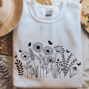 K&ouml;nnte beinhalten: Wei&szlig;es Sweatshirt mit einer schwarzen Linienzeichnung eines Blumenarrangements mit Schmetterlingen. Die Blumen umfassen G&auml;nsebl&uuml;mchen, Wildblumen und einen L&ouml;wenzahn.