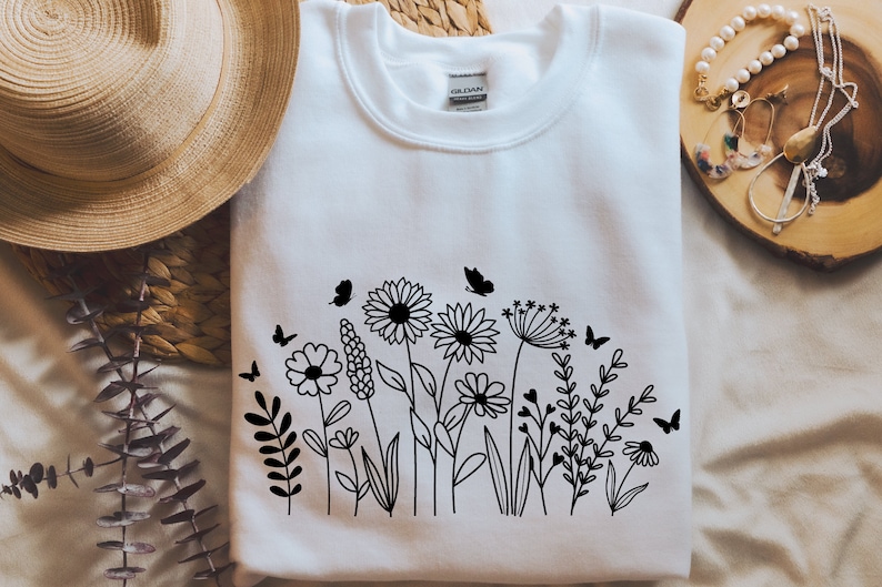 K&ouml;nnte beinhalten: Wei&szlig;es Sweatshirt mit einer schwarzen Linienzeichnung eines Blumenarrangements mit Schmetterlingen. Die Blumen umfassen G&auml;nsebl&uuml;mchen, Wildblumen und einen L&ouml;wenzahn.