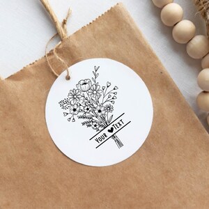 May include: A white, round tag with a black floral bouquet design and the text "YOUR TEXT" is attached to a brown paper bag. The tag is tied with twine. Wooden beads are visible in the background.
