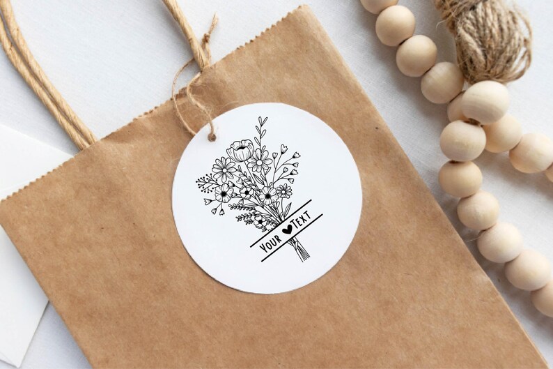 May include: A white, round tag with a black floral bouquet design and the text "YOUR TEXT" is attached to a brown paper bag. The tag is tied with twine. Wooden beads are visible in the background.