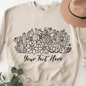 May include: A beige sweatshirt with a black and white graphic of cacti and succulents. The text "Your Text Here" is printed below the graphic.
