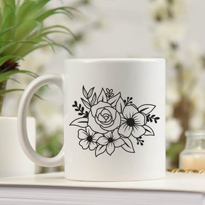 May include: White ceramic mug with a black floral design featuring a rose and other flowers. The mug has a handle and is sitting on a stack of white books.