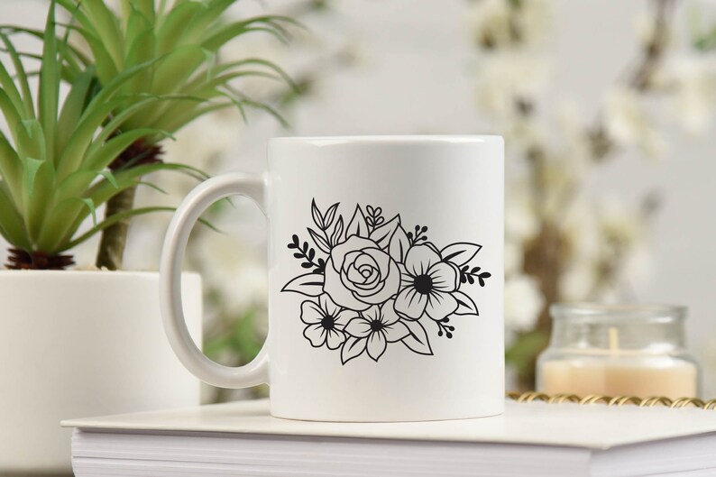 May include: White ceramic mug with a black floral design featuring a rose and other flowers. The mug has a handle and is sitting on a stack of white books.