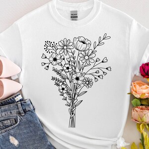 May include: White t-shirt featuring a black line drawing of a floral bouquet. The design includes various flowers and leaves tied together. The image also shows pink heels and blue jeans.