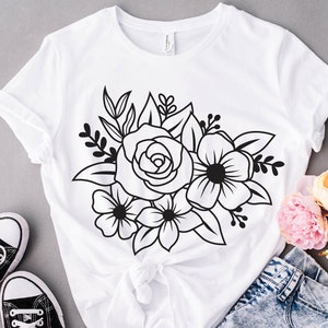 May include: A white t-shirt with a black floral design featuring a large rose and smaller flowers. The design is centered on the shirt and is surrounded by leaves.