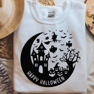 May include: White t-shirt with a black and white graphic design of a crescent moon, a haunted house, a witch flying on a broomstick, a tree, and pumpkins. The text "Happy Halloween" is written below the graphic.
