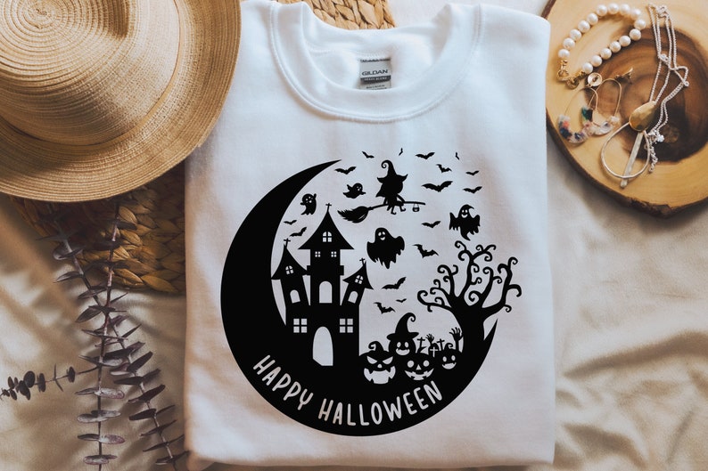 May include: White t-shirt with a black and white graphic design of a crescent moon, a haunted house, a witch flying on a broomstick, a tree, and pumpkins. The text "Happy Halloween" is written below the graphic.