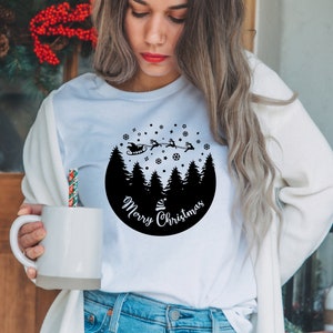 May include: White t-shirt with a black circle design featuring a silhouette of a snowy forest with Santa Claus flying overhead in his sleigh. The text "Merry Christmas" is written in a circle around the forest.