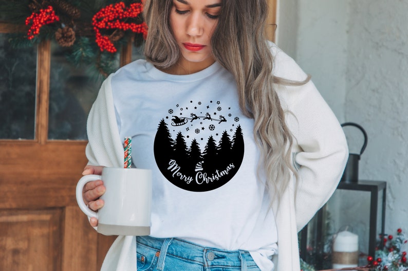 May include: White t-shirt with a black circle design featuring a silhouette of a snowy forest with Santa Claus flying overhead in his sleigh. The text "Merry Christmas" is written in a circle around the forest.