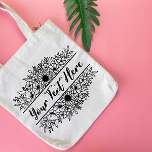 K&ouml;nnte beinhalten: Wei&szlig;e Canvas-Tragetasche mit schwarzem Blumendesign und dem Text "Your Text Here" auf der Vorderseite.