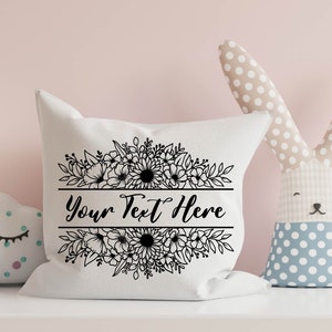 K&ouml;nnte beinhalten: Ein wei&szlig;es Kissen mit einem schwarzen Blumendesign und dem Text "Your Text Here" in einer dekorativen Schrift.