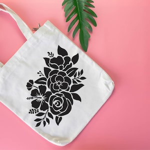 Pode incluir: Uma bolsa tote de lona branca com um design floral preto. A bolsa tem um fecho de correr e duas al&ccedil;as.