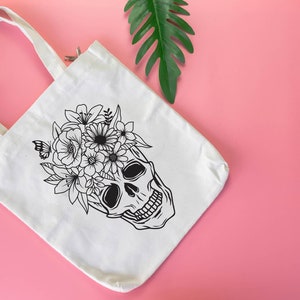 May include: White canvas tote bag with a black line drawing of a skull with flowers on top. The skull is smiling and has a floral crown.