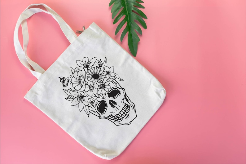 May include: White canvas tote bag with a black line drawing of a skull with flowers on top. The skull is smiling and has a floral crown.