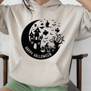 May include: A cream-colored hoodie with a black graphic of a crescent moon, a haunted house, a witch flying on a broomstick, ghosts, bats, and a tree. The text "Happy Halloween" is written in a circle around the graphic.