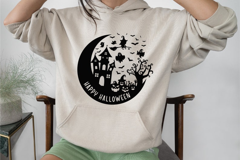 May include: A cream-colored hoodie with a black graphic of a crescent moon, a haunted house, a witch flying on a broomstick, ghosts, bats, and a tree. The text "Happy Halloween" is written in a circle around the graphic.