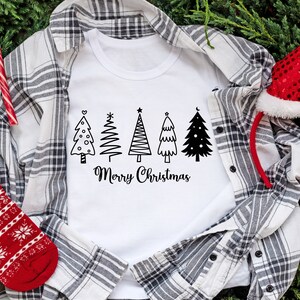 May include: White t-shirt with a black and white design of five Christmas trees and the text "Merry Christmas". The t-shirt is laid on a plaid shirt with a red and white candy cane and a red Santa hat.