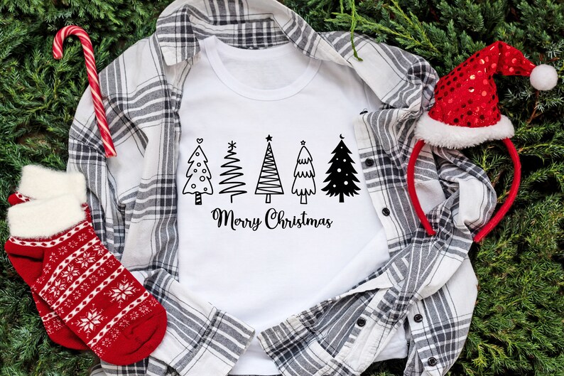 May include: White t-shirt with a black and white design of five Christmas trees and the text "Merry Christmas". The t-shirt is laid on a plaid shirt with a red and white candy cane and a red Santa hat.