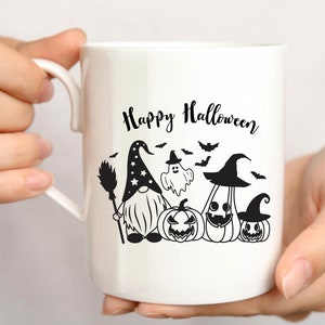 May include: White ceramic mug with a black and white design featuring a gnome, a ghost, and pumpkins. The text "Happy Halloween" is printed on the mug.