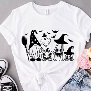 May include: White t-shirt with a black and white graphic of a gnome wearing a witch hat, a ghost, and three pumpkins. The gnome is holding a broom.