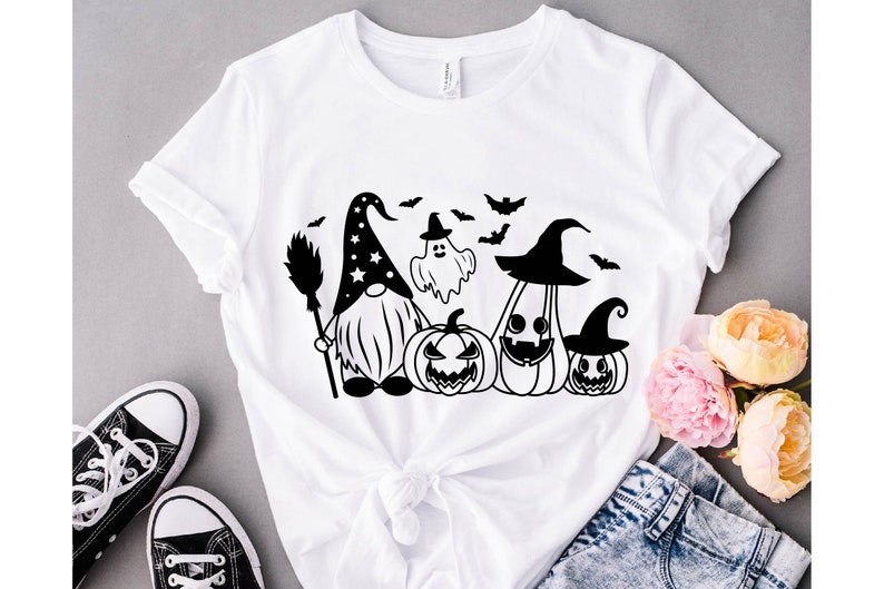May include: White t-shirt with a black and white graphic of a gnome wearing a witch hat, a ghost, and three pumpkins. The gnome is holding a broom.