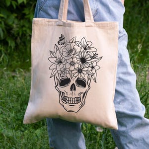 May include: A beige tote bag with a black and white illustration of a skull with flowers on top. The skull is wearing a floral crown.