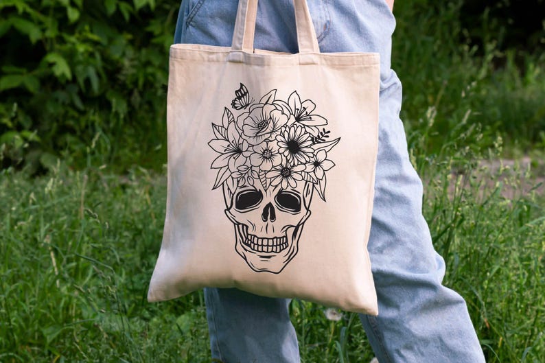 May include: A beige tote bag with a black and white illustration of a skull with flowers on top. The skull is wearing a floral crown.