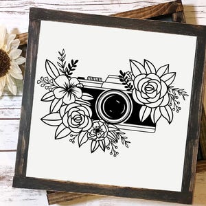K&ouml;nnte beinhalten: Schwarz-wei&szlig;-Illustration einer Kamera, die von Blumen umgeben ist. Die Kamera hat ein Objektiv und einen Riemen. Die Blumen sind stilisiert und haben ein einfaches Design.