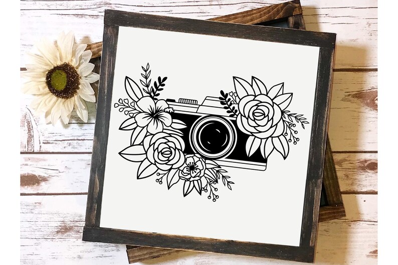 K&ouml;nnte beinhalten: Schwarz-wei&szlig;-Illustration einer Kamera, die von Blumen umgeben ist. Die Kamera hat ein Objektiv und einen Riemen. Die Blumen sind stilisiert und haben ein einfaches Design.