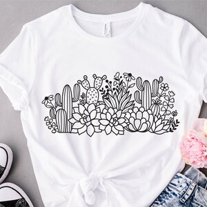 May include: A white t-shirt with a black line drawing of a variety of succulents and cacti. The design features a variety of shapes and sizes of succulents and cacti, as well as some small flowers.