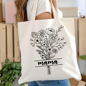 May include: Cream-colored tote bag featuring a black floral bouquet design with the word "mama" in a rectangular frame. The bag has long handles and is made of a sturdy fabric, suitable for carrying various items. The design is simple and elegant.
